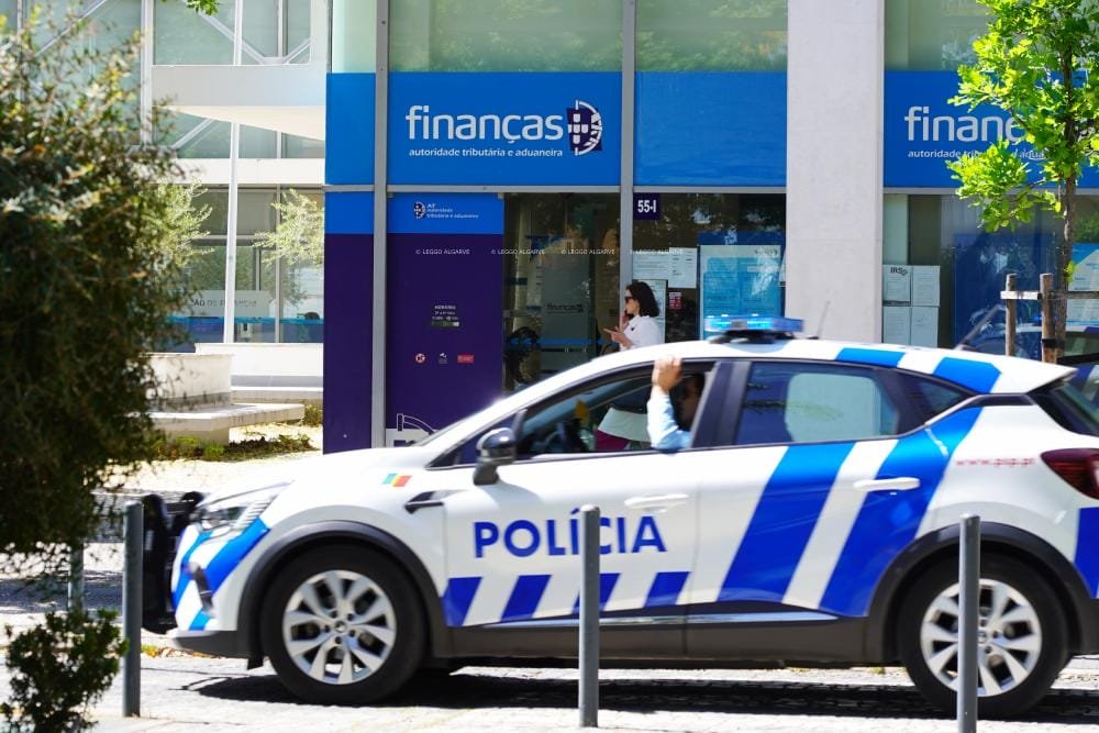 Un'auto della polizia blu e bianca è parcheggiata di fronte a un edificio per indagare sulle truffe alla Finanças tramite email. Il palazzo ha una facciata in vetro e la scritta "Finanças". L'edificio, contrassegnato da una segnaletica blu, funge da ufficio dell'autorità fiscale e doganale. Vicino all'ingresso c'è un individuo che passa. Alberi e dissuasori sono in primo piano.