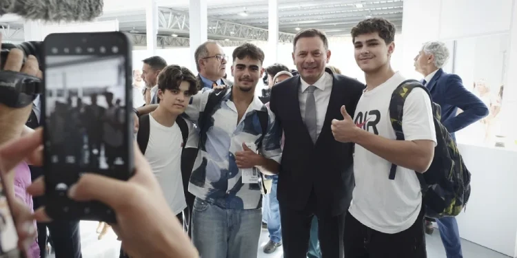 Un gruppo di tre giovani con Luis Montenegro, tutti sorridenti e che alzano il pollice, immagine inserita nel contesto della Legge di bilancio del Portogallo ( Orçamento de Estado ). La scena è stata catturata su uno smartphone da qualcuno in primo piano, in mezzo a un ambiente interno luminoso e moderno.