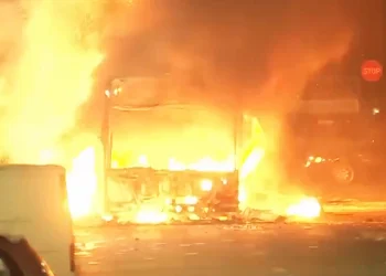Un autobus avvolto da fiamme luminose di notte, con un'intensa luce del fuoco che illumina l'area circostante, a Lisbona in una scena emblematica dei disordini urbani. Un segnale di stop è visibile sullo sfondo a destra, mentre detriti sono sparsi sul terreno.