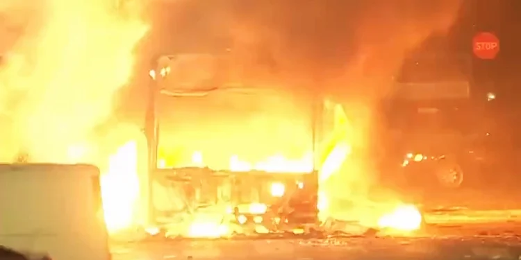 Un autobus avvolto da fiamme luminose di notte, con un'intensa luce del fuoco che illumina l'area circostante, a Lisbona in una scena emblematica dei disordini urbani. Un segnale di stop è visibile sullo sfondo a destra, mentre detriti sono sparsi sul terreno.