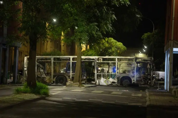 Nel cuore di una notte silenziosa, un autobus bruciato si erge come un duro promemoria dei disordini urbani di Lisbona. Circondato da alberi e illuminato dai lampioni, il veicolo gravemente danneggiato, con i suoi resti carbonizzati e i pannelli mancanti, si trova vicino agli edifici circostanti e a un attraversamento pedonale sullo sfondo.