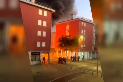 Un gruppo di persone è in piedi all'angolo di una strada a Lisbona, osservando un incendio in un edificio di mattoni rossi. Un denso fumo nero si solleva nel cielo, alludendo a possibili disordini urbani. La scena è scarsamente illuminata, il che suggerisce un mattino presto o una sera.