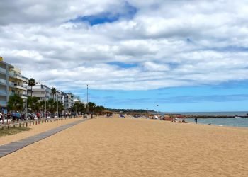 La spiaggia sabbiosa di Quarteira con persone che nonostante il meteo instabile si rilassano su questa spiaggia in Portogallo. Adiacente alla spiaggia c'è una passerella fiancheggiata da palme ed edifici moderni, che riflettono il soleggiato. Il cielo è parzialmente nuvoloso con una vista dell'oceano sullo sfondo.