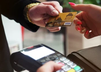 La mano di una persona che tiene una carta di credito Visa dorata per effettuare pagamenti elettronici in Portogallo. In basso, è visibile il terminale di pagamento, dove la carta sta per essere strisciata per una transazione MULTIBANCO.