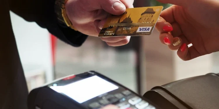 La mano di una persona che tiene una carta di credito Visa dorata per effettuare pagamenti elettronici in Portogallo. In basso, è visibile il terminale di pagamento, dove la carta sta per essere strisciata per una transazione MULTIBANCO.