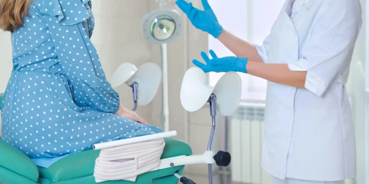 Una donna è seduta su un tavolo per visite mediche, di fronte alla ginecologa e probabilmente stanno discutendo della modalità dei parti cesarei nelle strutture sanitarie in Portogallo. Il medico indossa un camice bianco e guanti blu. L'ambiente sembra essere una clinica Privata.