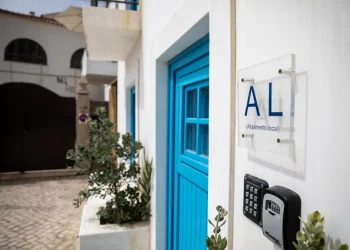 Un edificio bianco con una porta blu espone un cartello con restrizioni e la scritta "AL ( Alojamento Local )." È visibile un sistema di ingresso con tastiera, circondato da piante in vaso. La strada acciottolata e gli edifici bianchi adiacenti creano uno sfondo affascinante.