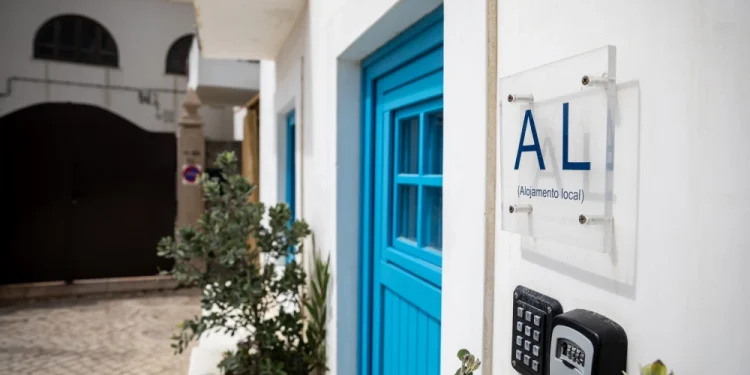 Un edificio bianco con una porta blu espone un cartello con restrizioni e la scritta "AL ( Alojamento Local )." È visibile un sistema di ingresso con tastiera, circondato da piante in vaso. La strada acciottolata e gli edifici bianchi adiacenti creano uno sfondo affascinante.