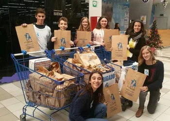 Un gruppo di sette volontari, sorridenti e con in mano borse per la raccolta del Banco Alimentar in Portogallo. Posano insieme in un centro commerciale e hanno riempito due carrelli della spesa con borse per una raccolta alimentare. Un piccolo albero di Natale è visibile sullo sfondo, aggiungendo un tocco festoso.