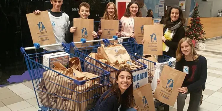 Un gruppo di sette volontari, sorridenti e con in mano borse per la raccolta del Banco Alimentar in Portogallo. Posano insieme in un centro commerciale e hanno riempito due carrelli della spesa con borse per una raccolta alimentare. Un piccolo albero di Natale è visibile sullo sfondo, aggiungendo un tocco festoso.