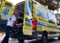 Un soccorritore sta caricando con cura un paziente su una barella in un'ambulanza gialla INEM Portogallo con le porte posteriori aperte. Il veicolo è parcheggiato su una strada acciottolata, incorniciata da alberi e un edificio sullo sfondo.