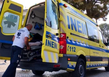 Un soccorritore sta caricando con cura un paziente su una barella in un'ambulanza gialla INEM Portogallo con le porte posteriori aperte. Il veicolo è parcheggiato su una strada acciottolata, incorniciata da alberi e un edificio sullo sfondo.