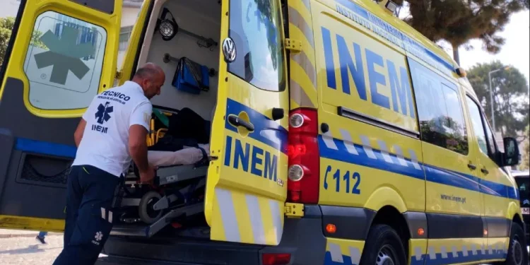 Un soccorritore sta caricando con cura un paziente su una barella in un'ambulanza gialla INEM Portogallo con le porte posteriori aperte. Il veicolo è parcheggiato su una strada acciottolata, incorniciata da alberi e un edificio sullo sfondo.