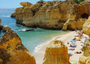 Una pittoresca spiaggia che richiama Turismo in Portogallo presenta scogliere dorate che circondano una stretta striscia di sabbia. I turisti prendono il sole e si rilassano sotto ombrelloni colorati vicino al mare turchese. Il cielo luminoso e la luce del sole esaltano la scena vibrante e serena, mostrando l'attrattiva turistica della regione.