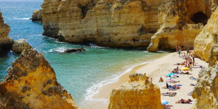 Una pittoresca spiaggia che richiama Turismo in Portogallo presenta scogliere dorate che circondano una stretta striscia di sabbia. I turisti prendono il sole e si rilassano sotto ombrelloni colorati vicino al mare turchese. Il cielo luminoso e la luce del sole esaltano la scena vibrante e serena, mostrando l'attrattiva turistica della regione.