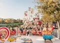 Si apre una scena festosa del parco divertimenti Wonderland Lisbona con una piccola ruota panoramica e una giostra a tema natalizio con figure di renne e pinguini. Molte persone si godono le attrazioni in una giornata di sole, mentre alberi e un'area erbosa offrono uno sfondo incantevole.