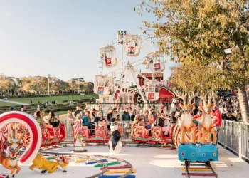 Si apre una scena festosa del parco divertimenti Wonderland Lisbona con una piccola ruota panoramica e una giostra a tema natalizio con figure di renne e pinguini. Molte persone si godono le attrazioni in una giornata di sole, mentre alberi e un'area erbosa offrono uno sfondo incantevole.