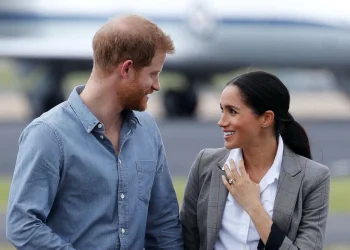 Harry e Meghan sono in piedi all'aperto, sorridenti l'uno all'altra. probabilmente appena atterrati in Portogallo. L'uomo indossa una camicia blu e la donna indossa un blazer grigio e una camicetta bianca. Un aereo è sfocato sullo sfondo.