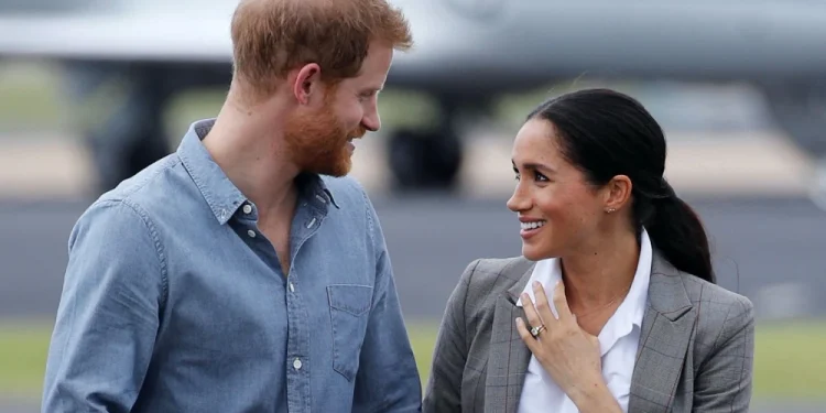 Harry e Meghan sono in piedi all'aperto, sorridenti l'uno all'altra. probabilmente appena atterrati in Portogallo. L'uomo indossa una camicia blu e la donna indossa un blazer grigio e una camicetta bianca. Un aereo è sfocato sullo sfondo.