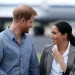 Harry e Meghan sono in piedi all'aperto, sorridenti l'uno all'altra. probabilmente appena atterrati in Portogallo. L'uomo indossa una camicia blu e la donna indossa un blazer grigio e una camicetta bianca. Un aereo è sfocato sullo sfondo.