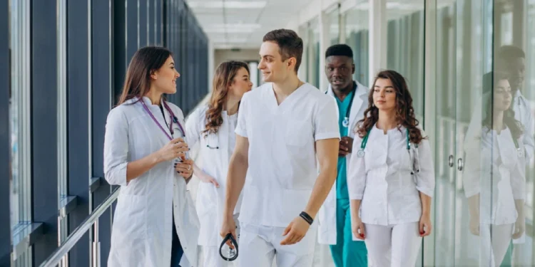 Un gruppo eterogeneo di professionisti sanitari, tra cui Medici Ucraini in camice bianco e camice, cammina lungo un corridoio dell'ospedale, impegnato in una conversazione e sorridente. Il corridoio è luminoso e rivestito di vetro, creando un'atmosfera moderna e accogliente che riflette l'integrazione.
