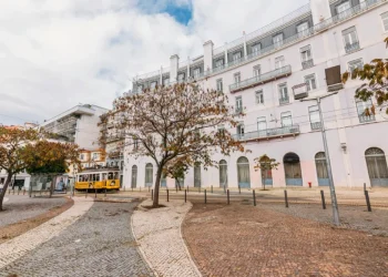 Riflettendo i mutevoli umori del meteo una strada acciottolata di Lisbona in Portogallo mostra un tram giallo che scivola davanti a un grande edificio bianco a più piani con finestre ad arco. Alberi spogli costeggiano il marciapiede sotto un cielo parzialmente nuvoloso.
