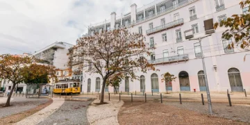 Riflettendo i mutevoli umori del meteo una strada acciottolata di Lisbona in Portogallo mostra un tram giallo che scivola davanti a un grande edificio bianco a più piani con finestre ad arco. Alberi spogli costeggiano il marciapiede sotto un cielo parzialmente nuvoloso.
