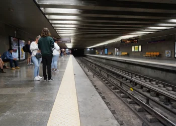 La piattaforma della Metro di Lisbona è ben illuminata, con solo poche persone in piedi e sedute durante lo sciopero. I binari attraversano il centro, fiancheggiati da pareti grigie adornate da manifesti pubblicitari, mentre i display in alto forniscono aggiornamenti.