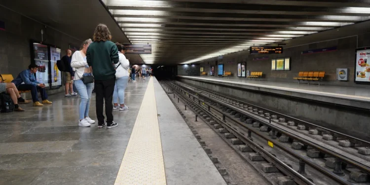 La piattaforma della Metro di Lisbona è ben illuminata, con solo poche persone in piedi e sedute durante lo sciopero. I binari attraversano il centro, fiancheggiati da pareti grigie adornate da manifesti pubblicitari, mentre i display in alto forniscono aggiornamenti.