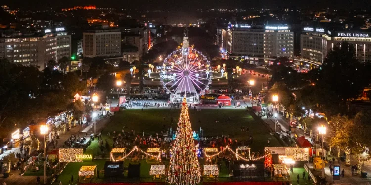 Una vivace scena notturna di Lisbona adornata con decorazioni natalizie, in questo periodo del Natale in Portogallo. Un grande albero di Natale illuminato si erge al centro, circondato da luci festive. Una ruota panoramica brilla sullo sfondo, in contrasto con il paesaggio urbano scuro.