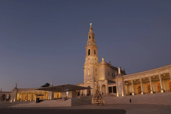 Una grande chiesa con una torre sormontata da una croce, illuminata al crepuscolo, si erge come una delle destinazioni affascinanti in Portogallo. Un grande albero di Natale adornato di luci abbellisce l'ampio cortile, fiancheggiato da colonnati ad arco, creando un'atmosfera natalizia serena e festosa.