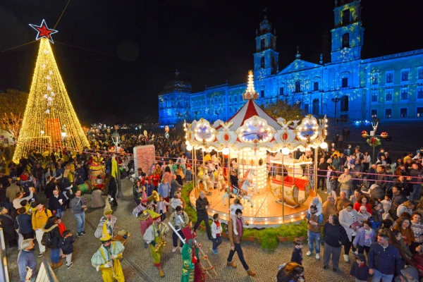 Una scena notturna di festa in Portogallo prende vita con una giostra illuminata, circondata da una folla vivace. Un grande albero di Natale illuminato si trova lì vicino e un edificio sullo sfondo si illumina di blu. Le persone si immergono nell'incantevole atmosfera del Natale in Portogallo.
