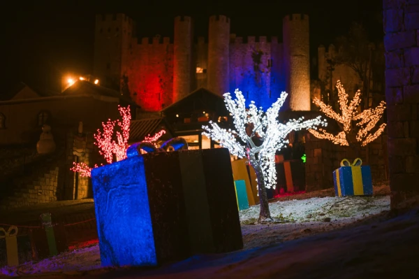 Una scena notturna con un castello illuminato sullo sfondo, illuminato di rosso e blu, cattura la magia del Natale in Portogallo. Grandi e colorate scatole regalo incartate e alberi adornati con luci bianche sono in primo piano, creando un'atmosfera festosa.