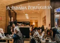 Le persone siedono ai tavoli della famosa catena di caffè A Padaria Portuguesa immerse in conversazioni o usando computer portatili. Una donna serve con grazia i clienti, esaltando l'atmosfera calda e invitante con il suo arredamento moderno.