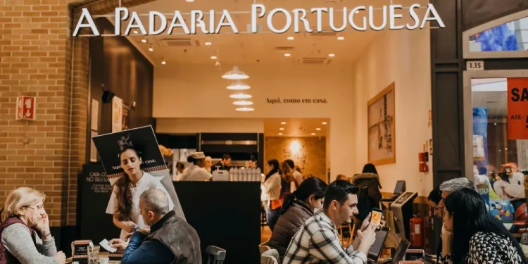 Le persone siedono ai tavoli della famosa catena di caffè A Padaria Portuguesa immerse in conversazioni o usando computer portatili. Una donna serve con grazia i clienti, esaltando l'atmosfera calda e invitante con il suo arredamento moderno.