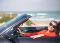 Una persona anziani con i capelli corti e gli occhiali da sole sorride seduta in un'auto decappottabile in riva al mare, forse assaporando la libertà ritrovata dopo aver deciso di rinnovare la patente. L' oceano e la spiaggia sabbiosa sono visibili sullo sfondo sotto un cielo terso, siamo in Algarve in Portogallo.