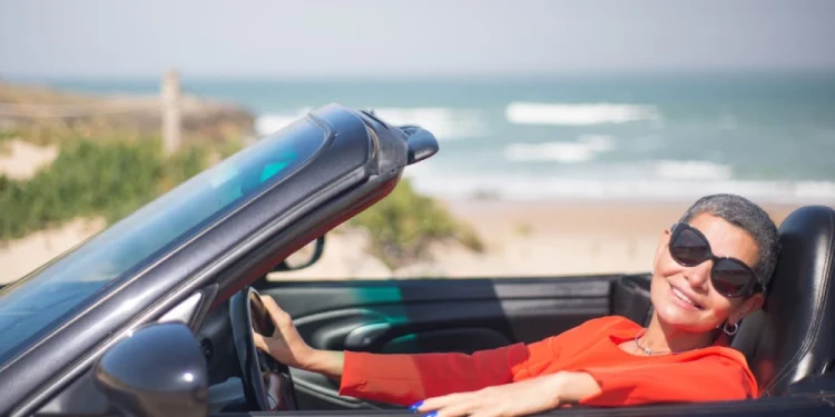 Una persona anziani con i capelli corti e gli occhiali da sole sorride seduta in un'auto decappottabile in riva al mare, forse assaporando la libertà ritrovata dopo aver deciso di rinnovare la patente. L' oceano e la spiaggia sabbiosa sono visibili sullo sfondo sotto un cielo terso, siamo in Algarve in Portogallo.
