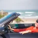 Una persona anziani con i capelli corti e gli occhiali da sole sorride seduta in un'auto decappottabile in riva al mare, forse assaporando la libertà ritrovata dopo aver deciso di rinnovare la patente. L' oceano e la spiaggia sabbiosa sono visibili sullo sfondo sotto un cielo terso, siamo in Algarve in Portogallo.