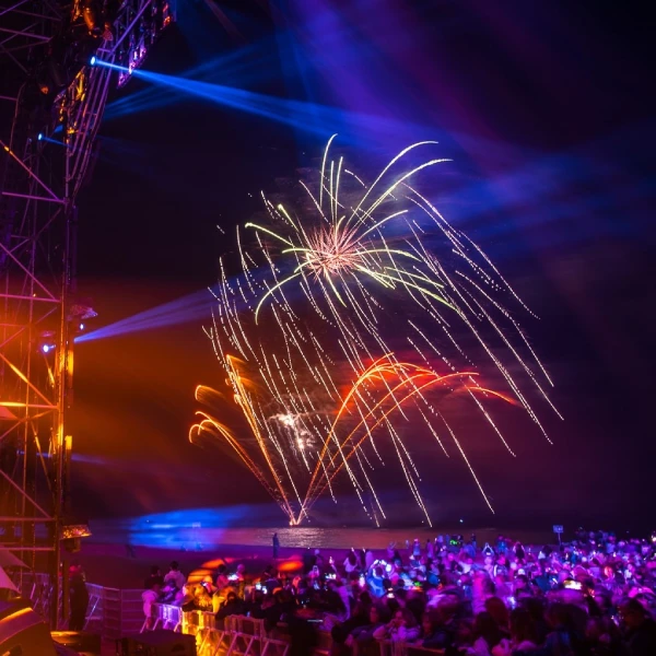 Fuochi d'artificio vibranti della festa di capodanno, illuminano il cielo notturno di Albufeira in Algarve su una folla all'aperto. Luci colorate e raggi dal palco creano un'atmosfera dinamica e festosa, mentre i festaioli del capodanno in spiaggia si godono i vivaci blu, viola e oro che contrastano magnificamente con lo sfondo scuro del mare.