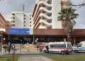 L'immagine raffigura la facciata dell'ospedale di Almada con un moderno edificio a più piani e scale che conducono all'ingresso, anche qui vi sono casi di cure sanitarie non pagate dagli stranieri. Un'ambulanza e diverse persone in cerca di cure sanitarie sono visibili vicino all'ingresso, mentre una palma si erge sul lato destro.