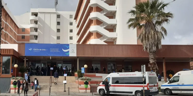L'immagine raffigura la facciata dell'ospedale di Almada con un moderno edificio a più piani e scale che conducono all'ingresso, anche qui vi sono casi di cure sanitarie non pagate dagli stranieri. Un'ambulanza e diverse persone in cerca di cure sanitarie sono visibili vicino all'ingresso, mentre una palma si erge sul lato destro.