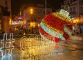 Una scena di strada festosa di notte a simboleggiare gli eventi di Natale a Coimbra presenta un grande ornamento natalizio illuminato con strisce rosse, bianche e dorate. L'hashtag "#COIMBRA" brilla accanto. Gli edifici circostanti e le luci natalizie completano questo incantevole sfondo di eventi.