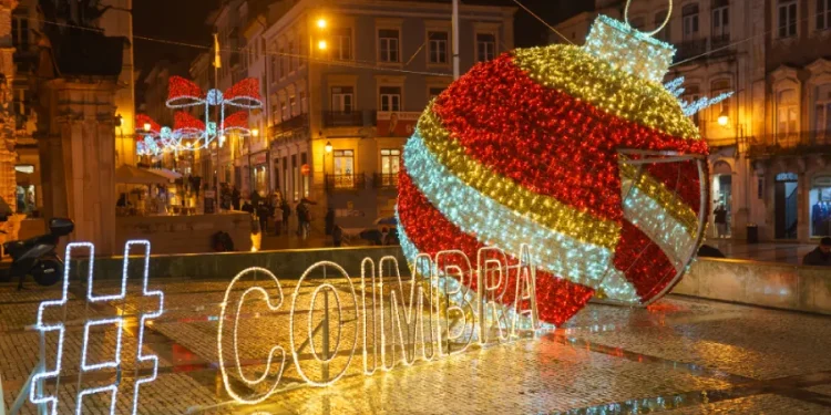Una scena di strada festosa di notte a simboleggiare gli eventi di Natale a Coimbra presenta un grande ornamento natalizio illuminato con strisce rosse, bianche e dorate. L'hashtag "#COIMBRA" brilla accanto. Gli edifici circostanti e le luci natalizie completano questo incantevole sfondo di eventi.