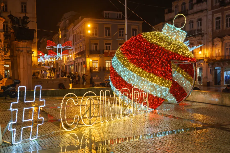 Una scena di strada festosa di notte a simboleggiare gli eventi di Natale a Coimbra presenta un grande ornamento natalizio illuminato con strisce rosse, bianche e dorate. L'hashtag "#COIMBRA" brilla accanto. Gli edifici circostanti e le luci natalizie completano questo incantevole sfondo di eventi.