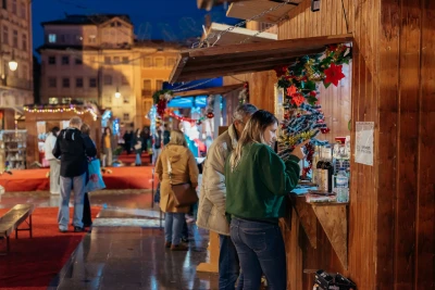 Un'accogliente scena notturna in un mercato all'aperto di Coimbra presenta bancarelle di legno. Le persone curiosano e si godono l'atmosfera natalizia festosa, con luci e decorazioni che aggiungono calore all'ambiente. Alcune persone sono radunate in una bancarella, affascinate dagli affascinanti oggetti esposti.
