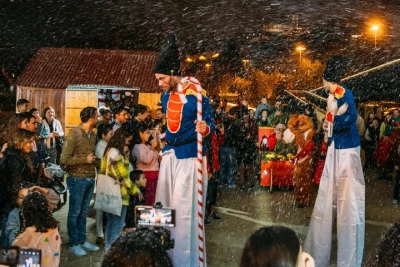 La gente si diverte a un evento festoso all'aperto a Coimbra di notte con la neve artificiale che cade. Due artisti sui trampoli vestiti da soldatini giocattolo intrattengono la folla, che comprende bambini e adulti. Sono visibili una bancarella di legno e decorazioni natalizie.