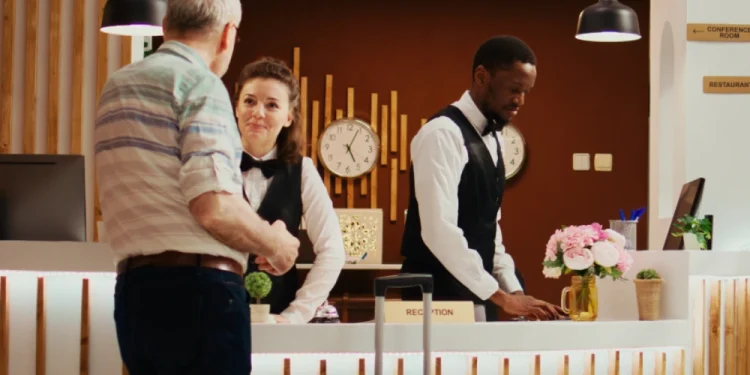 L' importanza dei lavoratori stranieri in Portogallo anche alla reception di un hotel che presenta due membri dello staff, una donna e un uomo in uniforme, che aiutano un ospite con una valigia. Dietro di loro, gli orologi mostrano diversi fusi orari e i cartelli evidenziano vari comfort.