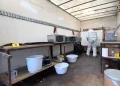 L'interno del laboratorio di cocaina smantellato in Portogallo una persona con equipaggiamento protettivo lavora vicino a diversi forni a microonde su un tavolo. Secchi e contenitori sono sul pavimento e vari oggetti sono sparsi sugli scaffali. Sono visibili i marcatori gialli delle prove.