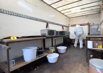 L'interno del laboratorio di cocaina smantellato in Portogallo una persona con equipaggiamento protettivo lavora vicino a diversi forni a microonde su un tavolo. Secchi e contenitori sono sul pavimento e vari oggetti sono sparsi sugli scaffali. Sono visibili i marcatori gialli delle prove.