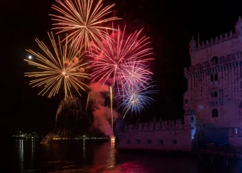 Con un meteo favorevole, nel cielo i fuochi d'artificio colorati scoppiano sulla torre di Belem vicino al fiume Tejo nella notte di Capodanno a Lisbona in Portogallo. Il cielo è illuminato da rossi, arancioni e blu brillanti, che si riflettono nell'acqua calma sottostante, creando una scena abbagliante.
